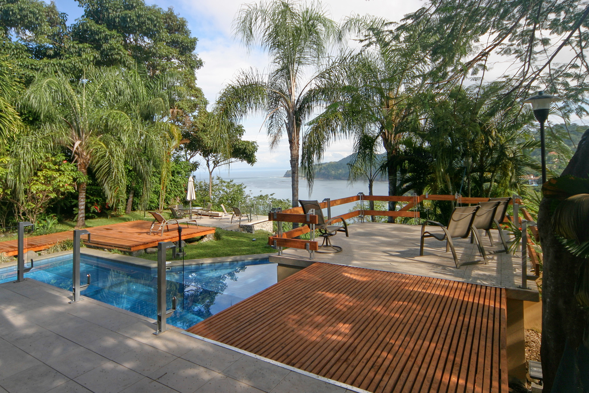 Casa-Beer-Fuscaldo_Playa-Escondida Terrace view of mountains transitioning to the Pacific Ocean, with a section of a pool, wooden deck, and bone porcelanato pathways visible
