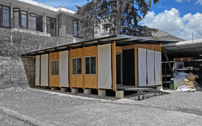 Built view of Taller Huella Colectiva, a compact wood-framed workshop with cantilevered metal roof, raised on concrete pads, with white tarpaulin panels alternating across the façade between vertical wood cladding and window openings. Surrounding context is desaturated to highlight the workshop's material composition.