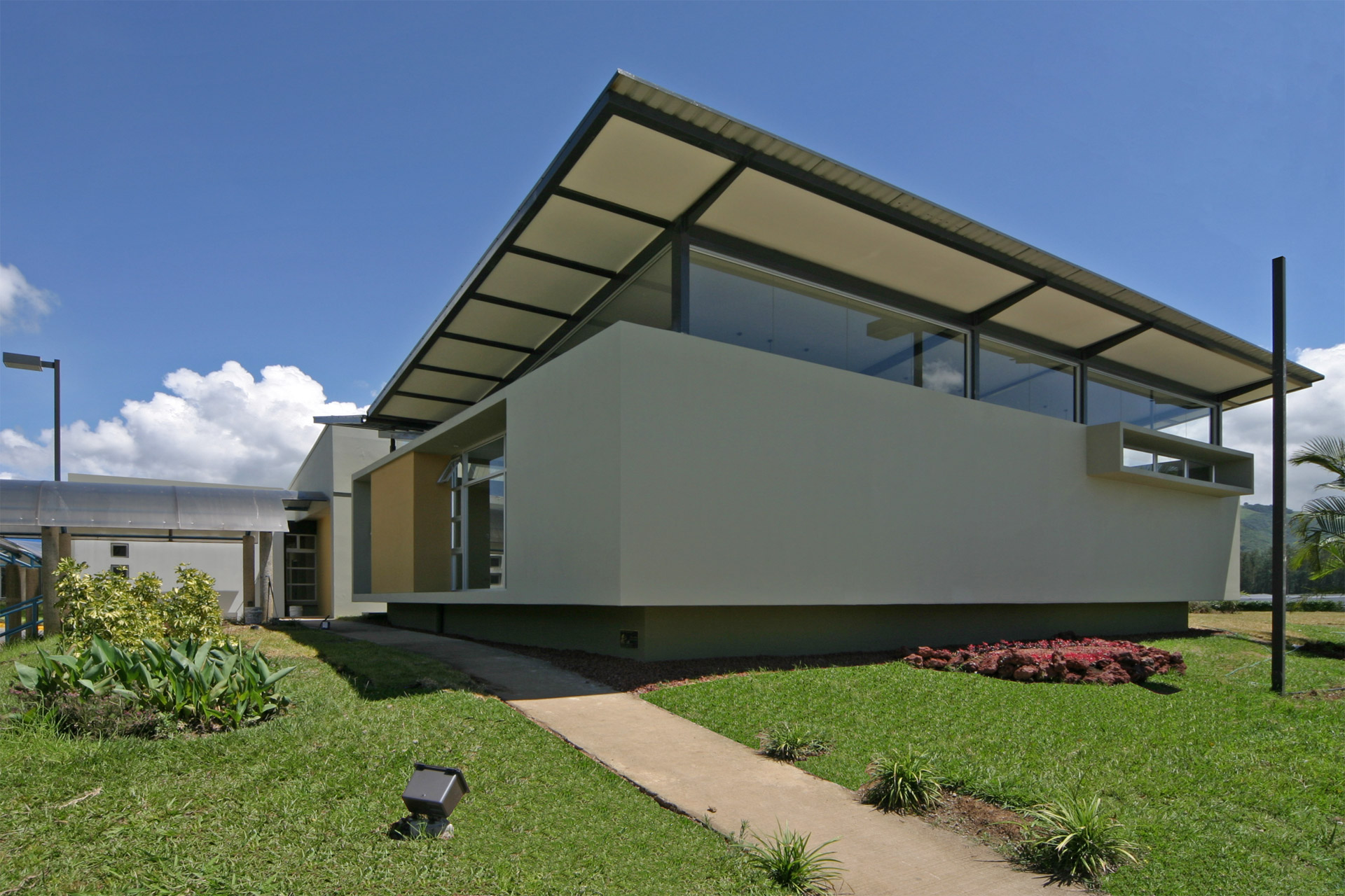 Outdoor view of the Novartis Software Development Office, surrounded by lush green grass. The corner of the building is featured with a distinctive butterfly roof, accented in green-gray and yellow. A sunny day showcases the architectural elegance of the structure. Costa Rican tropical design