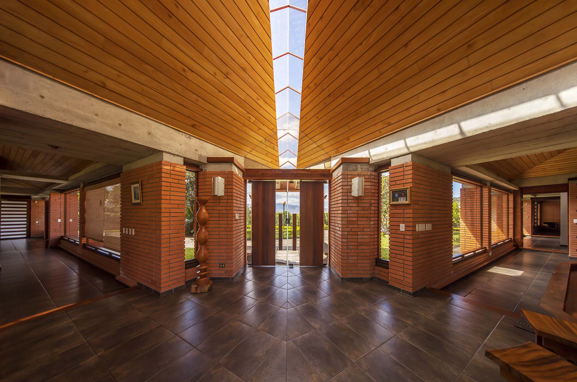 ymmetrical view from within the Soto House lobby, looking towards the main entrance door, with extended views down the wing hallways. The entrance doors, a mix of tempered glass and solid wood, appear as a floating wood panel under the skylight. Notable features include heavy brick columns, wooden ceilings, and concrete beams - a showcase of Costa Rican architecture