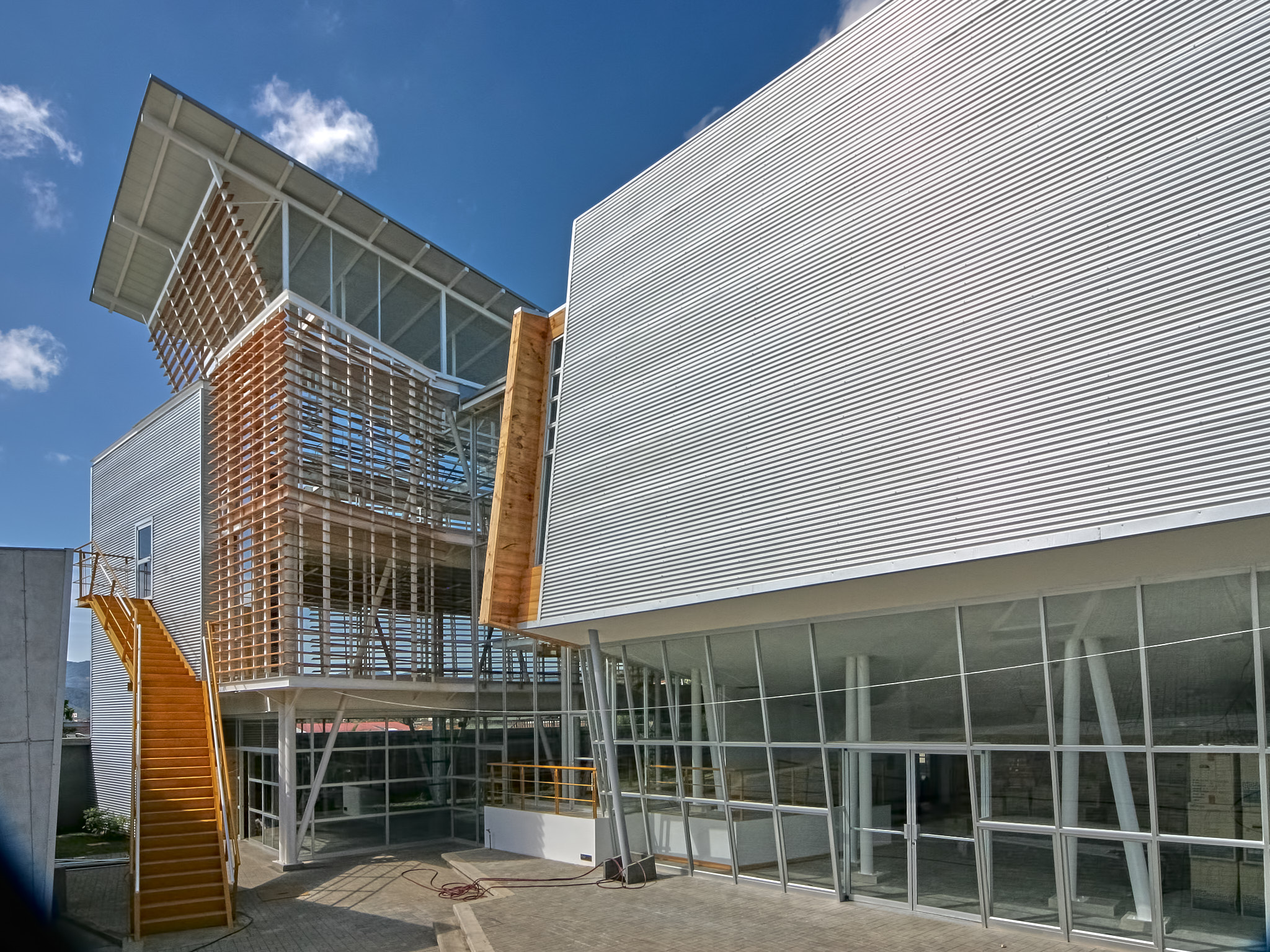 East facade of Veritas University, showing the glass gallery, central glass atrium entrance, and the yellow exterior steel stair of the workshop building