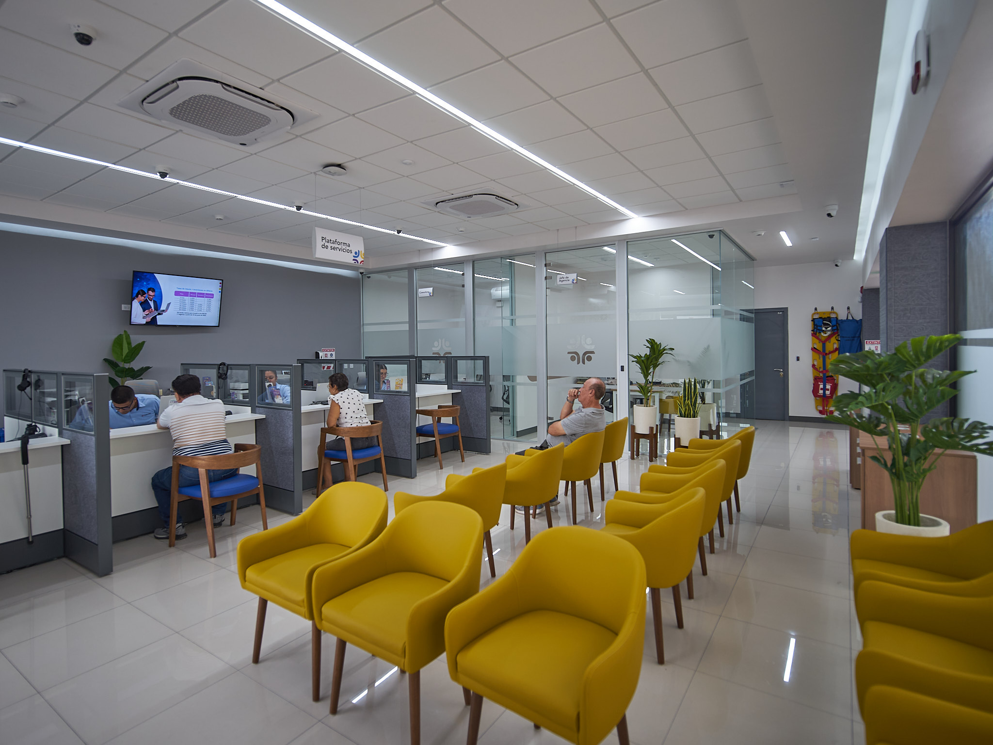 Vista del área de espera con las plataformas de atención a un lado y el cubo de vidrio del coworking al otro. Waiting area with service platforms on one side and a glass-enclosed coworking space on the other.